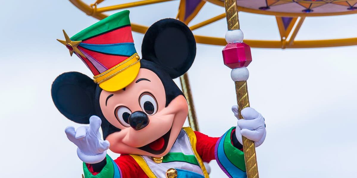 Mickey Mouse, in a colorful outfit and striped hat, waves cheerfully while standing on a parade float adorned with vibrant decorations. The background is a clear blue sky.