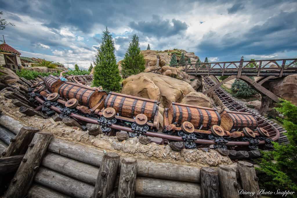 The Seven Dwarfs Mine Train twists through rugged rocks, lush trees, and a bridge beneath clouds at Magic Kingdom—pure Disney thrills!.