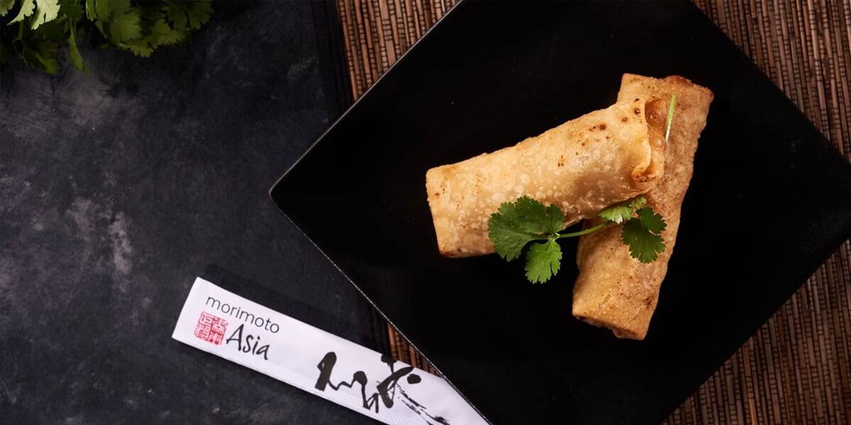 Two golden-brown spring rolls garnished with cilantro are served on a black plate with Morimoto Asia chopsticks—one of the Best Disney Springs Restaurants—set on a dark surface with a woven mat and fresh cilantro in the corner.