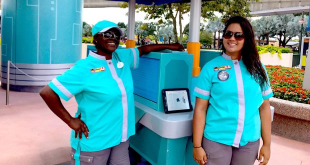 Two staff members in teal and gray uniforms with name badges stand and smile outdoors at a theme park, next to a teal kiosk with a tablet on it. There are trees, flowers, and a walkway in the background.