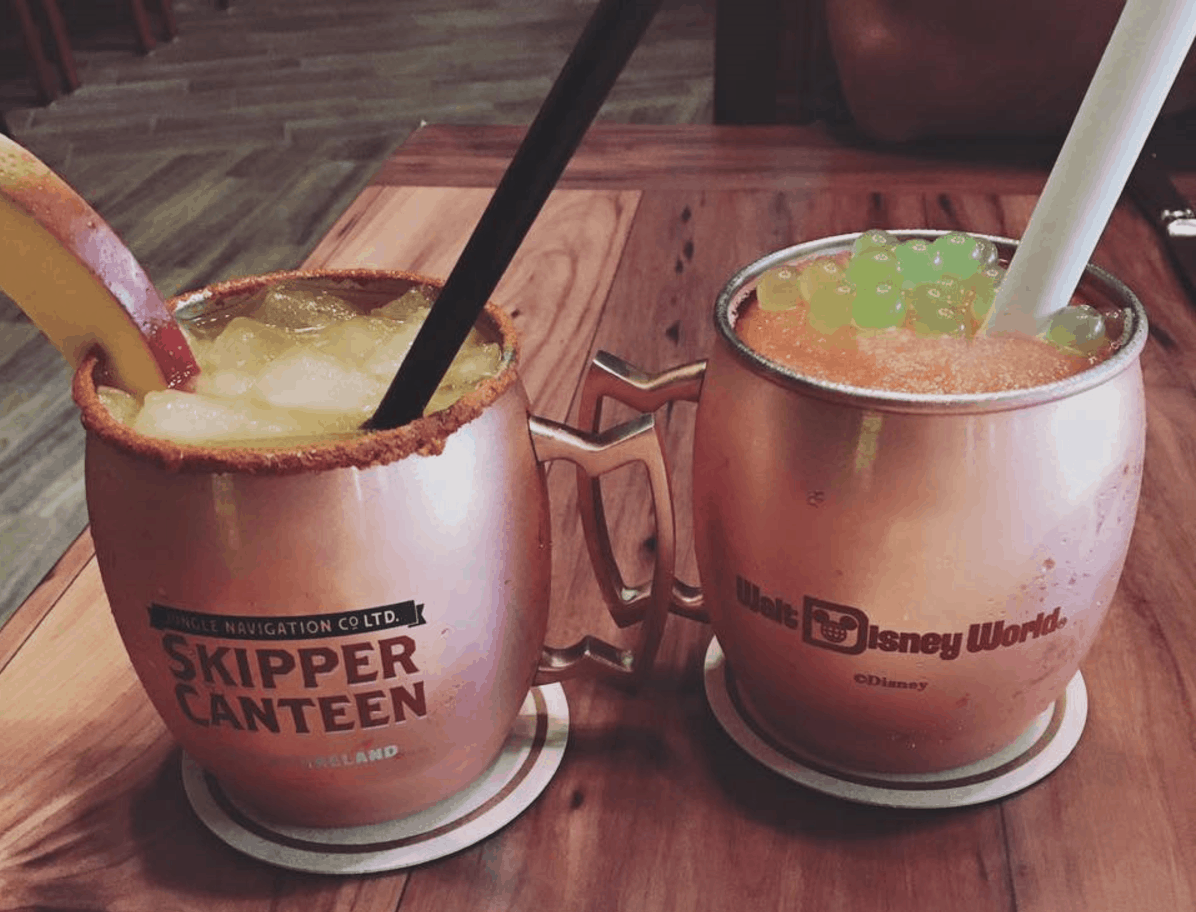 Copper mugs with cocktails sit on a wooden table, sporting "Skipper Canteen" and "Walt Disney World" logos for Disney fans.