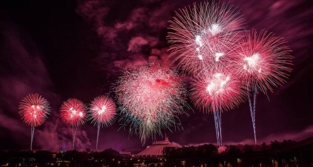 Vivid pink and white fireworks burst above Cinderella Castle and lush trees, capturing the enchanting spectacle of a Disney World night.