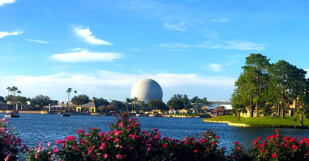 A large, white geodesic sphere rises in the distance behind trees, flowers, and a lake under a blue sky at Epcot in Walt Disney World, Florida.