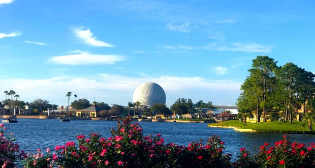 A large, white geodesic sphere rises in the distance behind trees, flowers, and a lake under a blue sky at Epcot in Walt Disney World, Florida.