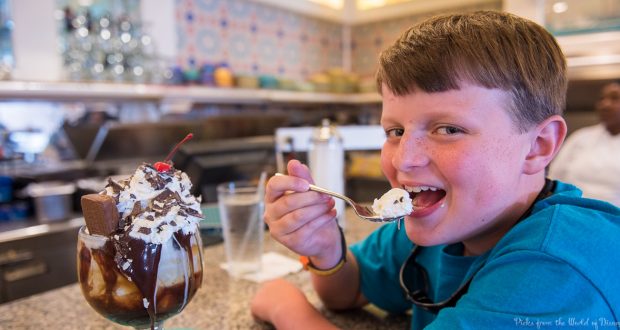 Smiling boy savoring a massive ice cream sundae topped with whipped cream, chocolate, and a cherry in a Disney World-style diner.