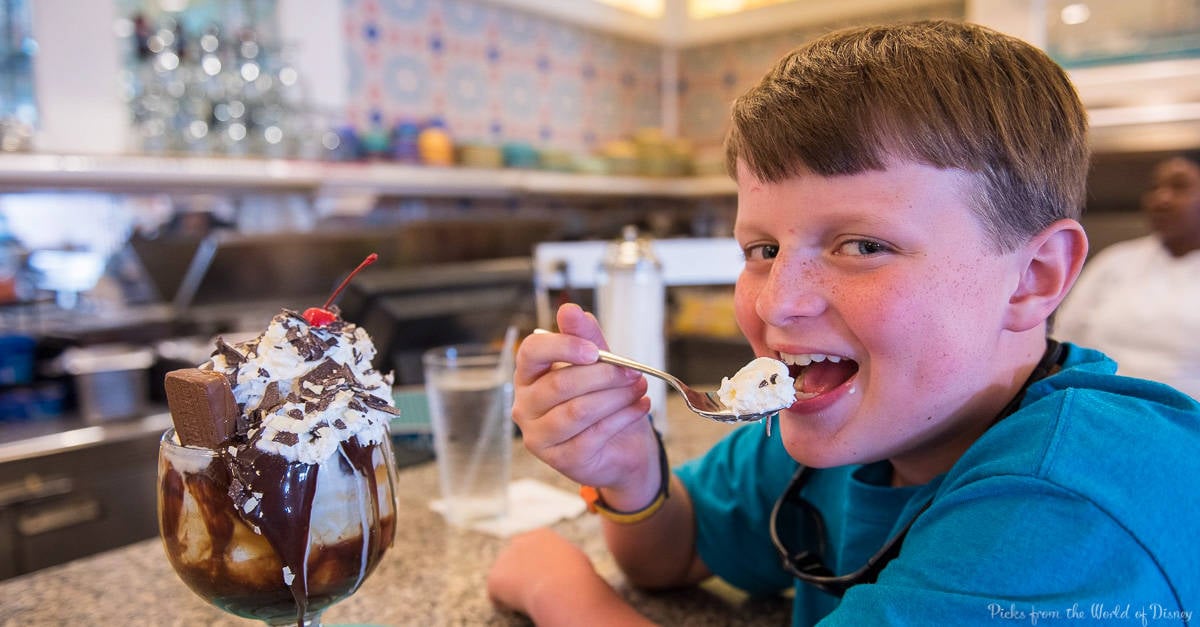 Young guest in a blue shirt beams while digging into a classic Disney World sundae topped with whipped cream, chocolate, and cherry.