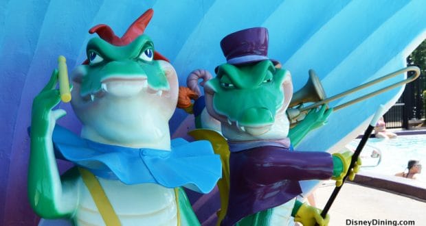 Alligator statues, one sporting a top hat and trombone, stand by the pool at Disney’s Moderate Resort against a blue backdrop.