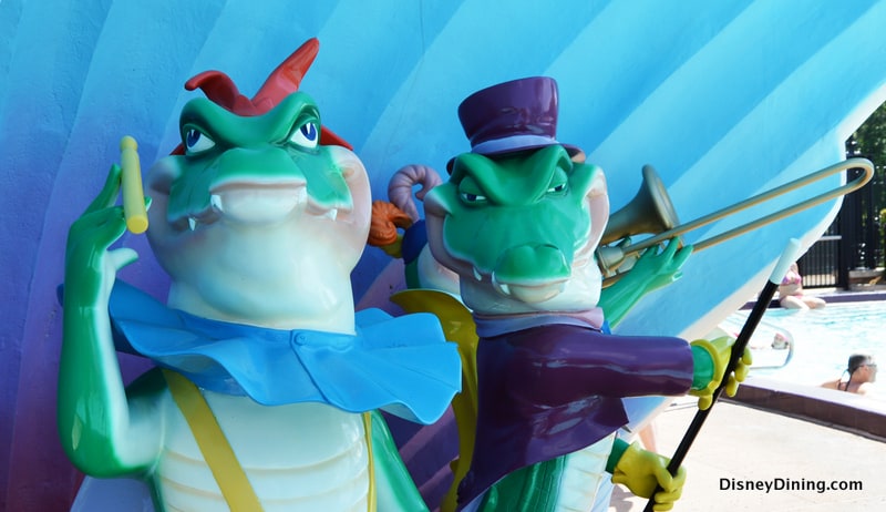 Alligator statues, one sporting a top hat and trombone, stand by the pool at Disney’s Moderate Resort against a blue backdrop.