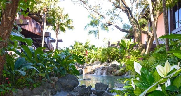 Tropical oasis with a small waterfall and pond, lush greenery evoking Disney’s Polynesian Village Resort near the hotel buildings.