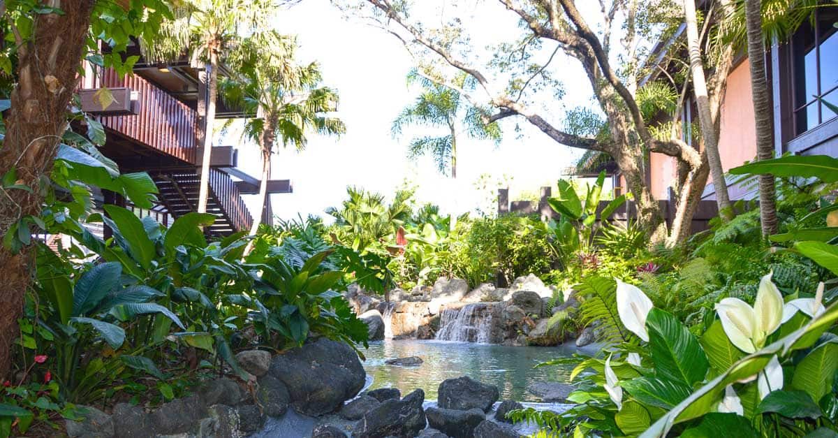 Tropical oasis with a small waterfall and pond, lush greenery evoking Disney’s Polynesian Village Resort near the hotel buildings.