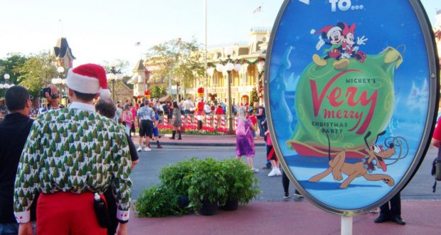 A guest in holiday gear poses by Mickey’s Very Merry Christmas Party sign at Disney World’s entrance, ready for festive park secrets.