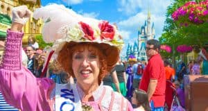 Disney character in a pink outfit and flowered hat greets guests, with Cinderella Castle and crowds at Walt Disney World, Orlando.