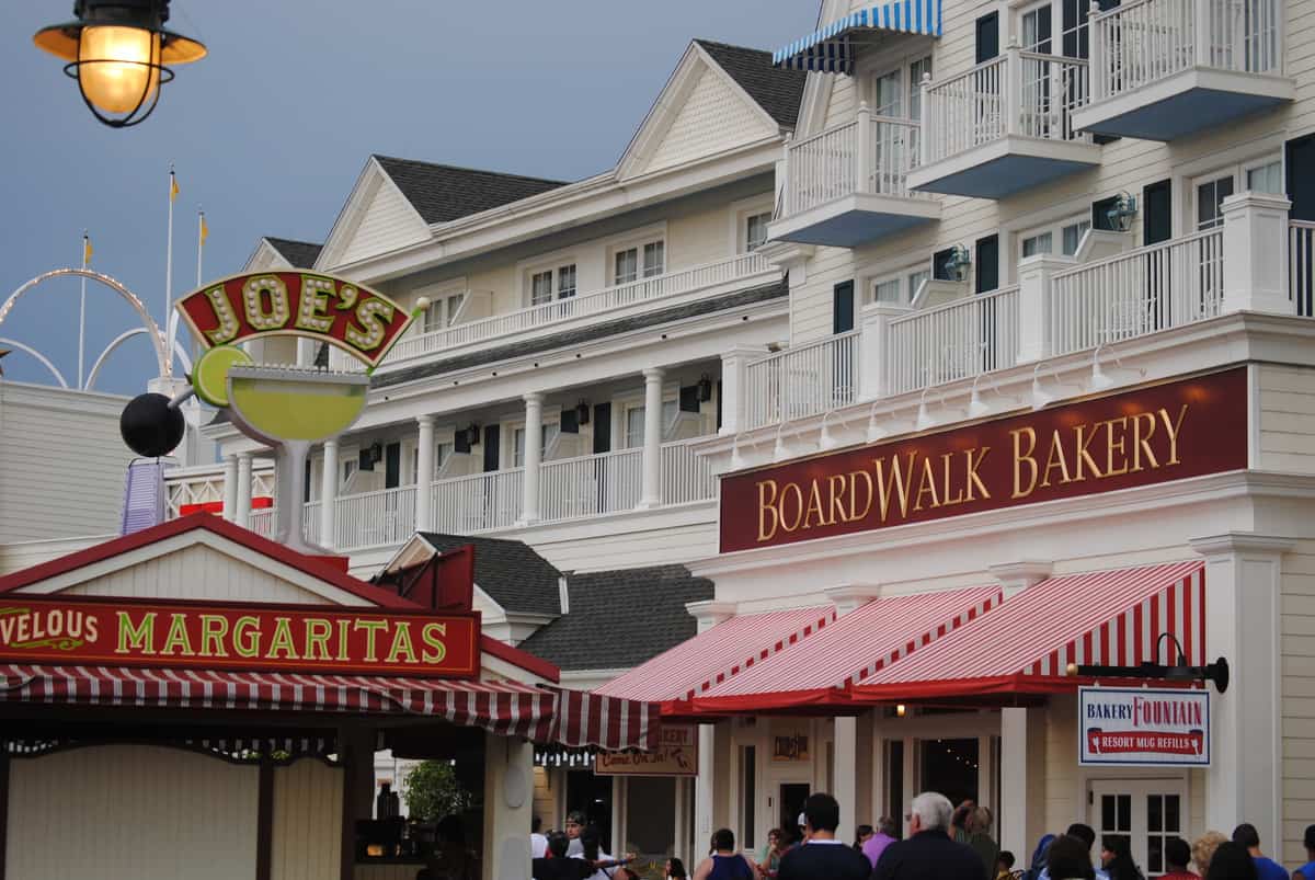 Guests stroll past Boardwalk Bakery and Joe’s Margaritas on Disney's lively Boardwalk, surrounded by charming white buildings.