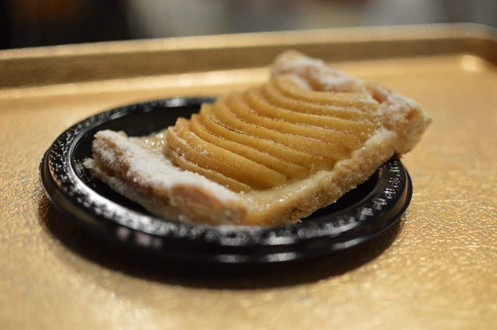 A slice of apple tart topped with powdered sugar at Les Halles in EPCOT, served on a black plate over a gold tray.