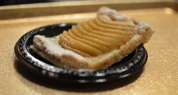 A slice of apple tart topped with powdered sugar at Les Halles in EPCOT, served on a black plate over a gold tray.