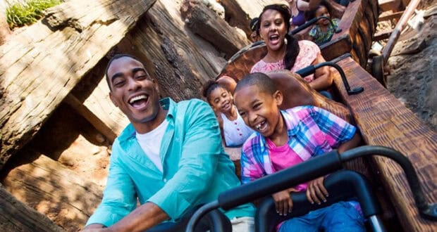 A family of four, smiling and laughing, rides a wooden roller coaster together on a sunny day. With Walt Disney World tips in mind, they make the most of the thrill, the father and son in front and mother and daughter right behind.