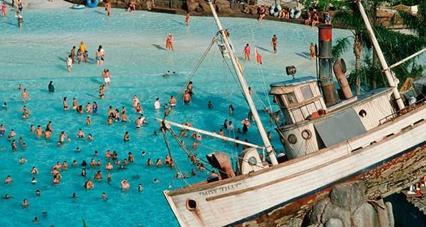 Lively pool scene with swimmers and a tilted pirate ship on rocks, evoking classic Disney water parks and adventure-themed resorts.
