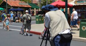 A photographer with a tripod and camera captures a scene on a sunny day in a theme park. People walk by, including a man pushing a stroller. Shops and umbrellas are visible in the background.
