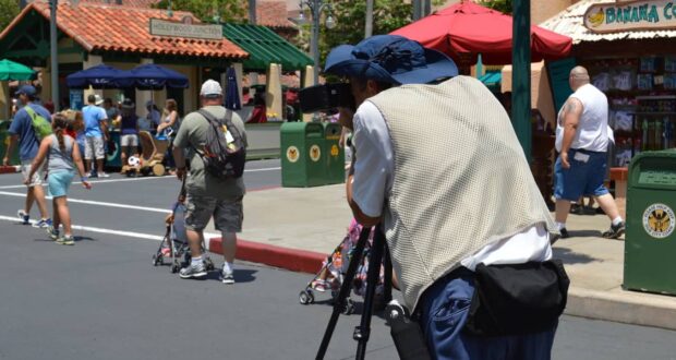 A photographer with a tripod and camera captures a scene on a sunny day in a theme park. People walk by, including a man pushing a stroller. Shops and umbrellas are visible in the background.