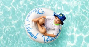 A young boy wearing a blue hat and white shirt smiles while sitting in a white inflatable tube floating in a clear, blue swimming pool, reminiscent of fun moments at Disney Water Parks.