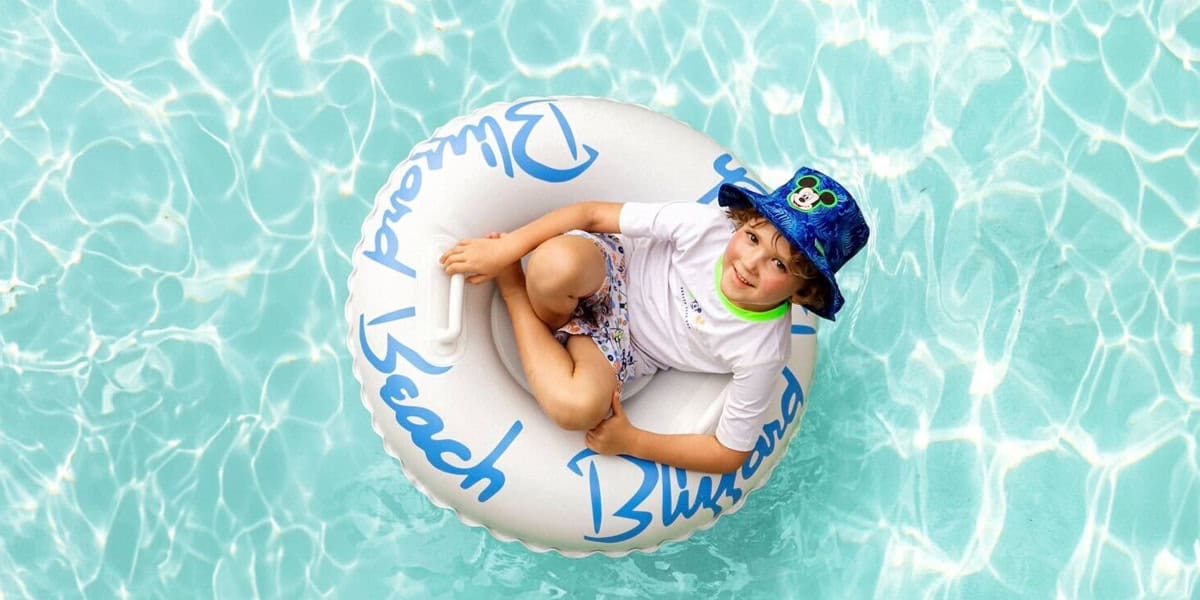 A young boy wearing a blue hat and white shirt smiles while sitting in a white inflatable tube floating in a clear, blue swimming pool, reminiscent of fun moments at Disney Water Parks.