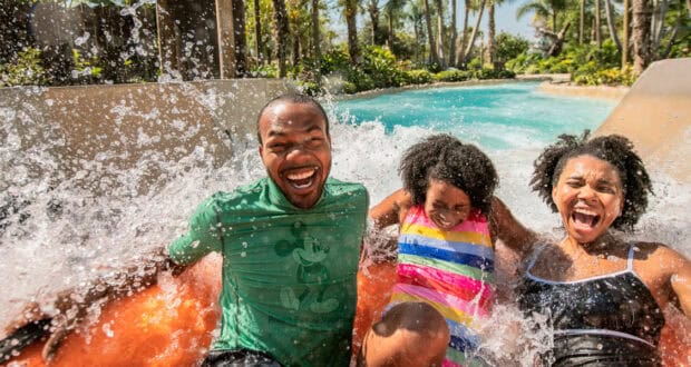 Three people are laughing and getting splashed with water while riding an orange raft at a Disney water park, surrounded by palm trees and greenery under a sunny sky—one of many fun Disney Water Parks facts to discover.