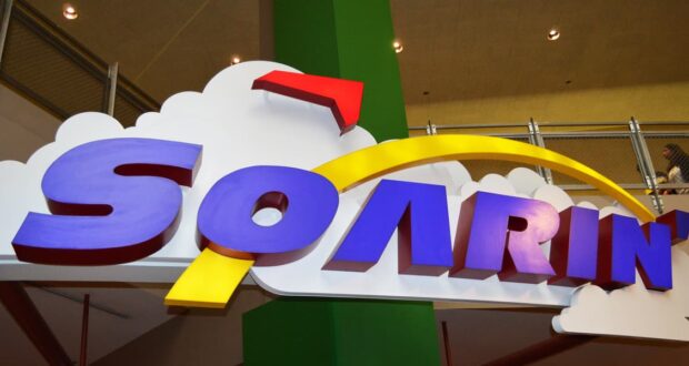 Soarin’ Around the World" sign with clouds and yellow arc, indoors by railing and green pillar—pure Disney Parks adventure vibes!.