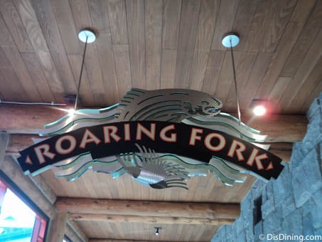 Roaring Fork sign with a fish motif hangs under rustic wooden beams, inviting guests to dine at Disney’s Wilderness Lodge.
