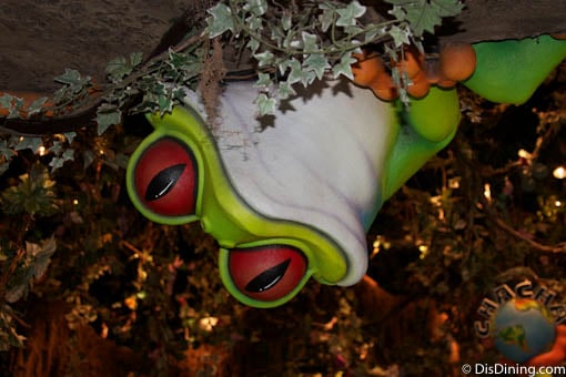 A vibrant cartoon frog with red eyes playfully hangs upside down in lush vines, adding kid-friendly fun to Disney Springs.