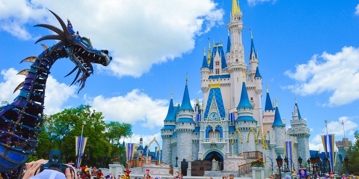 A large metal dragon float passes in front of Cinderella’s Castle at Disney World during a parade, with crowds of people gathered under a bright blue sky with white clouds.