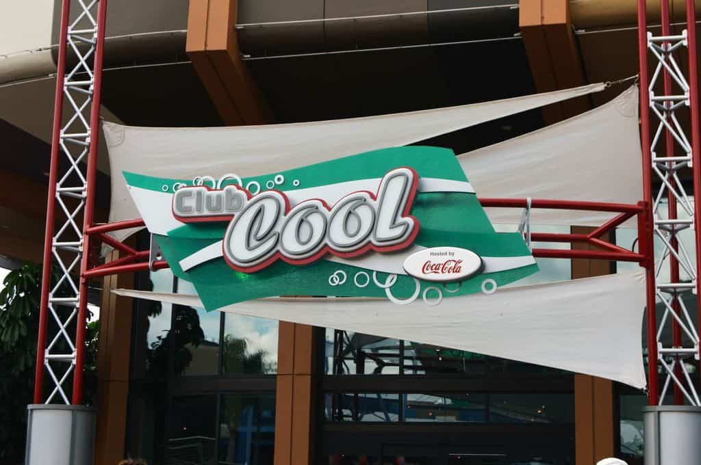 Club Cool’s green and white sign with Coca-Cola logo welcomes guests at Epcot, framed by bold red poles and festive fabric.