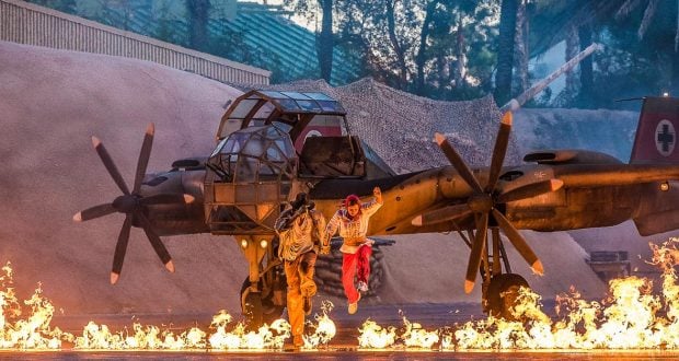 Guests watch as stunt actors dash from fiery explosions beside a classic military plane, recreating iconic Hollywood action scenes.