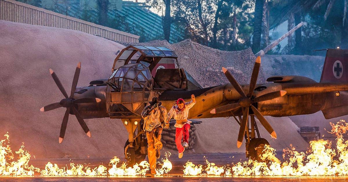 Guests watch as stunt actors dash from fiery explosions beside a classic military plane, recreating iconic Hollywood action scenes.