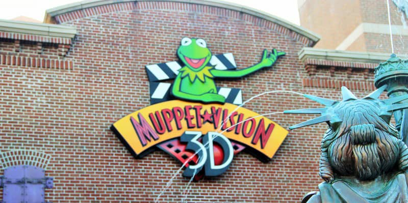 A colorful "Muppet Vision 3D" sign featuring Kermit the Frog is displayed on a brick building. In the foreground, part of a fountain statue resembling the Statue of Liberty is visible.