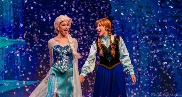 Elsa and Anna perform on stage at a Disney park, singing as snowflake confetti falls, bringing Frozen magic to the audience.
