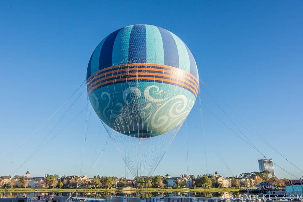 A large, colorful hot air balloon with blue and orange stripes is tethered to the ground with cables, floating above buildings under a clear blue sky.