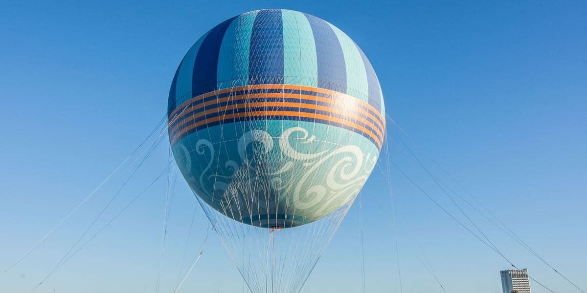 A large, colorful hot air balloon with blue and orange stripes is tethered to the ground with cables, floating above buildings under a clear blue sky.