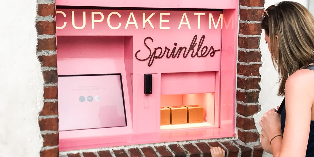 A woman stands in front of a pink Sprinkles cupcake ATM built into a brick wall. Three cupcake boxes are visible inside the machine’s open window.