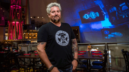 A stylish guest with spiked hair and tattoos inside Planet Hollywood Observatory, vibrant lights shining above empty dining tables.