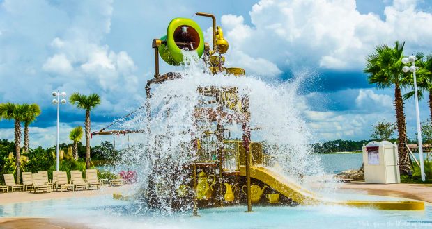 At a sunny Disney World water park, a giant bucket drenches the vibrant play area, palm trees, and loungers all around.