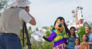A photographer captures a smiling family posing with Goofy at an amusement park, with water slides and trees in the background—it's a perfect Memory Maker moment.