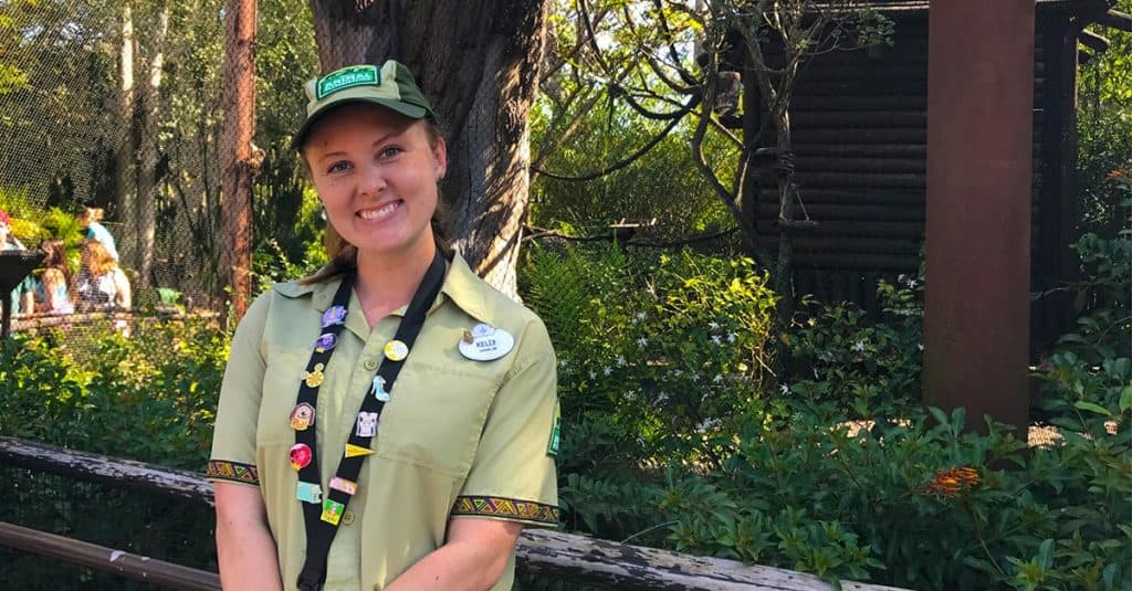 A cheerful Cast Member in classic green attire greets guests under lush trees at a Disney park, ready to assist with magic.