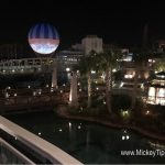 Disney Springs at night with a glowing bridge, soaring striped balloon, palm trees, and Planet Hollywood shining in the background.