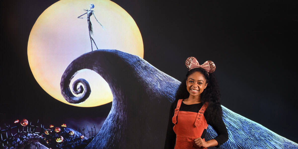 A smiling woman wearing Minnie Mouse ears and an orange overall dress stands in front of a Memory Maker backdrop featuring Jack Skellington from "The Nightmare Before Christmas" silhouetted against a large moon.