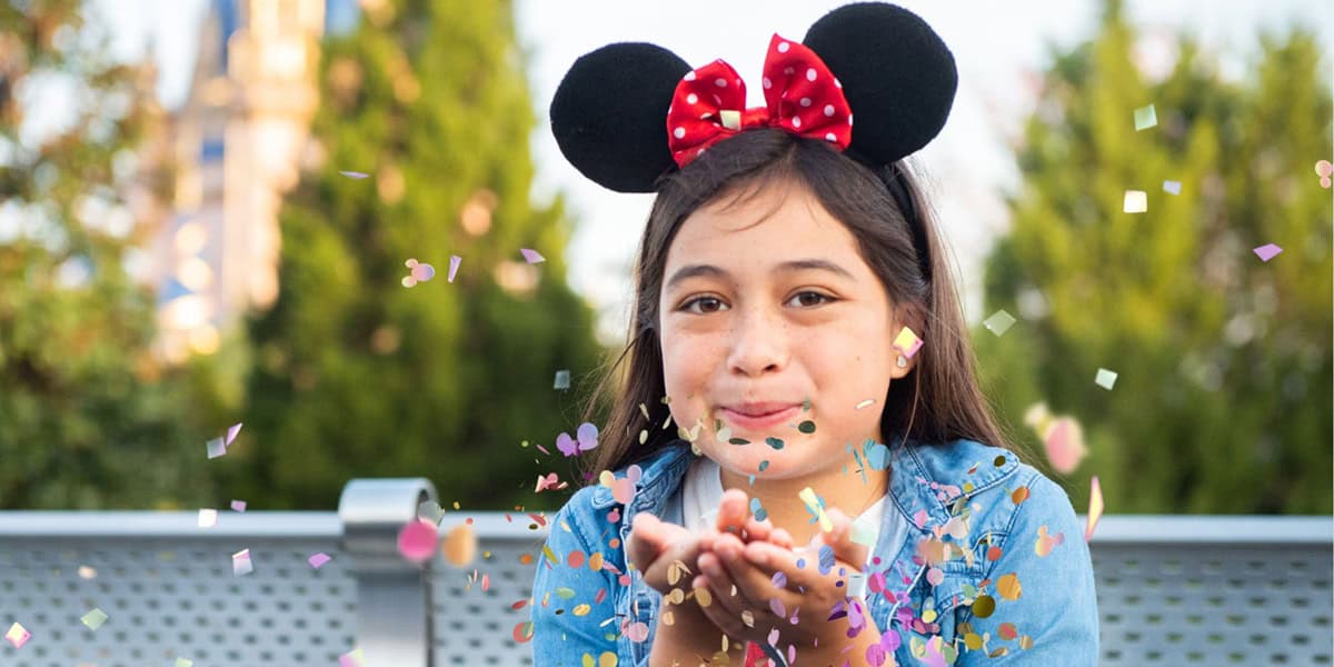 A young girl wearing Minnie Mouse ears and a red bow blows colorful confetti from her hands while smiling outdoors, capturing a Memory Maker moment. Green trees and a blurred castle are visible in the background.