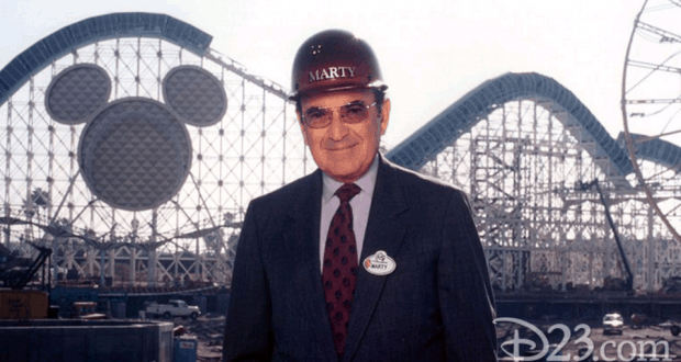 Disney Imagineer in suit and hard hat stands by a coaster, Mickey Mouse silhouette behind him at Walt Disney World—classic Sklar vibe.