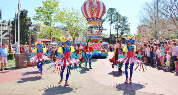 Disney parade performers in vibrant costumes dance past a colorful hot air balloon float at Magic Kingdom.