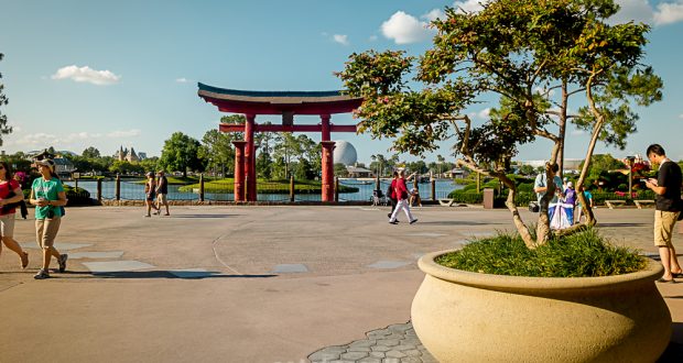 Guests stroll by a grand potted tree and iconic red Torii gate beside the Epcot lake on a bright, sunny Disney day.