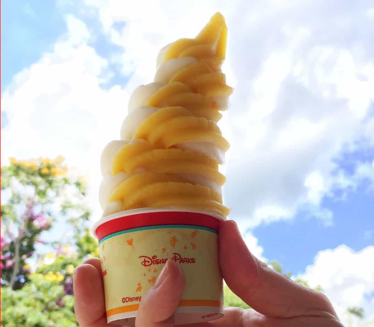 A refreshing Dole Whip swirl of pineapple and vanilla soft serve in a Disney Parks cup, held up to the magical blue sky.