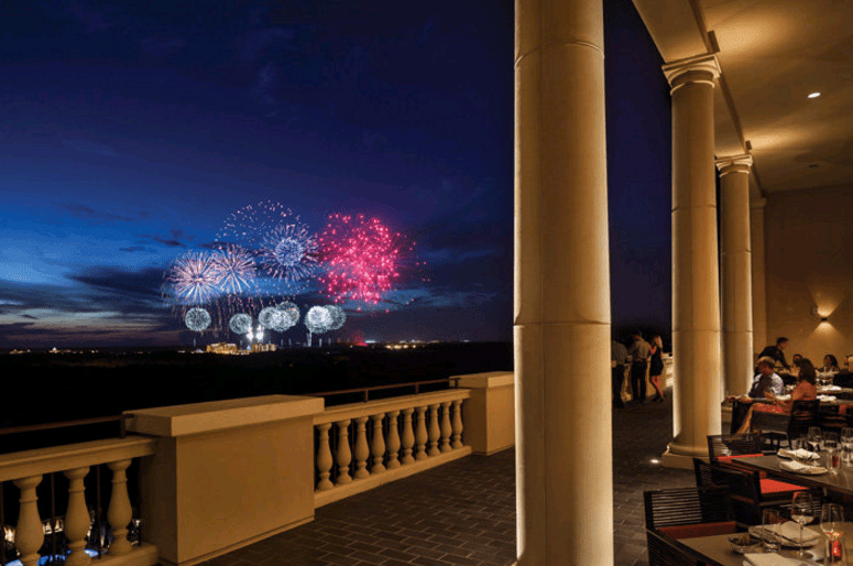 Guests savor dinner on an open-air terrace as vibrant fireworks light up the sky, capturing the magic of a Walt Disney World night.